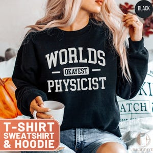 May include: Black sweatshirt with the text "WORLDS OKAYEST PHYSICIST" in white block letters. The sweatshirt is part of a t-shirt, sweatshirt, and hoodie collection. The word "BLACK" is in a black circle in the upper right corner.