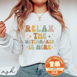 May include: A light gray sweatshirt with the text "RELAX THE WATCHMAKER IS HERE" in retro-style lettering. The design includes orange, yellow, green, and pink colors. A circular graphic at the bottom right indicates color and design options.