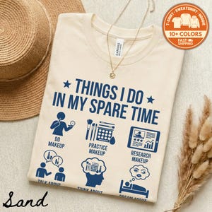 May include: A sand-colored t-shirt with the text "THINGS I DO IN MY SPARE TIME" and makeup-related icons in navy blue. The shirt also has the words "DO MAKEUP", "PRACTICE MAKEUP", and "RESEARCH MAKEUP".