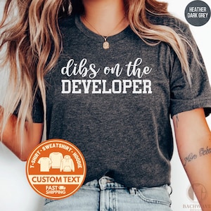 May include: Heather dark grey t-shirt with the text "dibs on the DEVELOPER" in white. The shirt has a crew neck and short sleeves. A custom text option is available. The shirt is part of a t-shirt, sweatshirt, and hoodie collection.