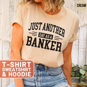 May include: Cream-colored t-shirt with the text "JUST ANOTHER DAY AS A BANKER" in black block letters. The shirt also has the words "T-SHIRT SWEATSHIRT & HOODIE" in a red box. The model is wearing blue jeans.
