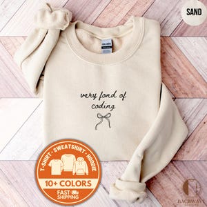 May include: Sand-colored sweatshirt with the text "very fond of coding" and a bow graphic. The sweatshirt has a crew neck and long sleeves. A circular graphic shows the options for t-shirt, sweatshirt, and hoodie.