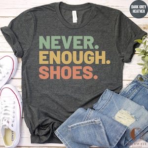May include: A dark grey heather t-shirt with the text "NEVER. ENOUGH. SHOES." printed in a retro style with green, yellow, and orange colors.