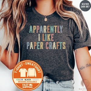 May include: Heather dark grey t-shirt with the text "APPARENTLY I LIKE PAPER CRAFTS" in pastel colors. The shirt also has a graphic indicating the availability of black and white designs. The shirt is a t-shirt or sweatshirt.