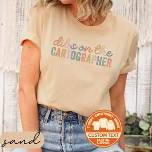 May include: Sand-colored t-shirt with rolled sleeves featuring the text "dibs on the CARTOGRAPHER" in pastel colors. The shirt is paired with blue jeans. A circular graphic at the bottom indicates options for custom text on a t-shirt, sweatshirt, or hoodie.