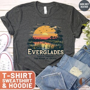 May include: Dark grey heather t-shirt with a vintage-style illustration of a sunset over a river with the text "Everglades - Paddle Through The River of Grass".