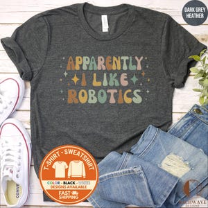 May include: A dark grey heather t-shirt featuring the text "APPARENTLY I LIKE ROBOTICS" in a retro, multi-colored font. The design includes small star accents in shades of orange, teal, and cream, printed on a short-sleeved crew neck shirt.