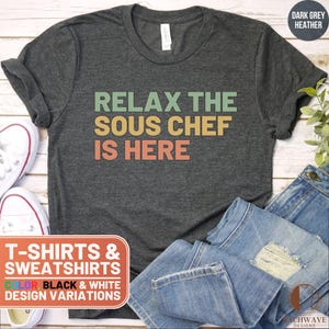 May include: Dark heather grey t-shirt with the text "Relax the Sous Chef is Here" in a retro font with green, yellow, and orange colors.
