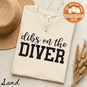 May include: Sand-colored t-shirt with the phrase "dibs on the DIVER" in black lettering. The shirt has a crew neck and is made of a soft material. A straw hat and a gold necklace are also visible.