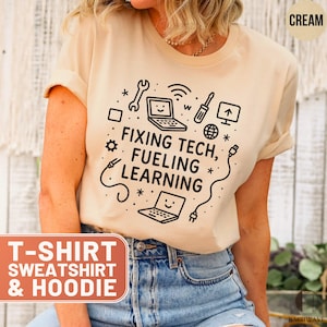 May include: Cream-colored t-shirt with the text "FIXING TECH, FUELING LEARNING" and tech-related graphics. The shirt also has the text "T-SHIRT SWEATSHIRT & HOODIE" in a rectangular box. The shirt is made of a soft material.