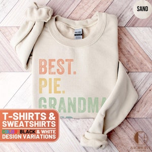 May include: A sand-colored sweatshirt with the text "BEST. PIE. GRANDMA" in a vintage style font. The text is in shades of orange, yellow, and green. The sweatshirt is available in black and white.
