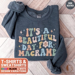 May include: Dark heather grey sweatshirt with the text "It's a beautiful day for macrame" in colorful retro font. The text is surrounded by small stars and a smiley face.