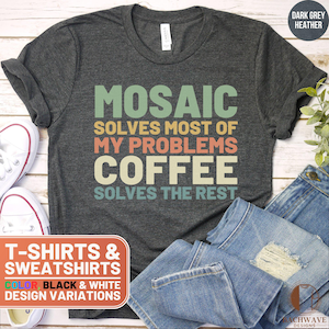 May include: A dark grey heather t-shirt with the text "Mosaic solves most of my problems coffee solves the rest" in a retro style font. The text is in shades of green, blue, orange, and brown. The t-shirt is displayed on a wooden surface with a pair of jeans and white sneakers.