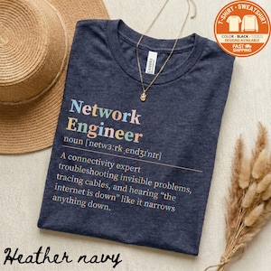 May include: Heather navy t-shirt with the text "Network Engineer" in colorful letters. The definition of a network engineer is printed below the title. A gold necklace and a straw hat are also in the image.