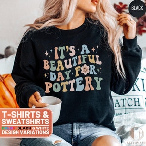 May include: Black sweatshirt with the text "It's a beautiful day for pottery" in colorful retro font. The text is surrounded by small stars and a flower with a smiley face.