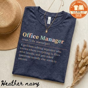 May include: Heather navy t-shirt with the words "Office Manager" in colorful text. The definition of "Office Manager" is printed below. A gold necklace and a straw hat are also in the image.