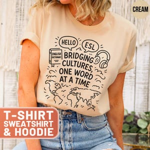May include: Cream-colored t-shirt with black graphic text: "HELLO ESL BRIDGING CULTURES, ONE WORD AT A TIME." The design includes a world map, headphones, and a dictionary. The bottom of the shirt has a rectangular text box that says "T-SHIRT SWEATSHIRT & HOODIE."
