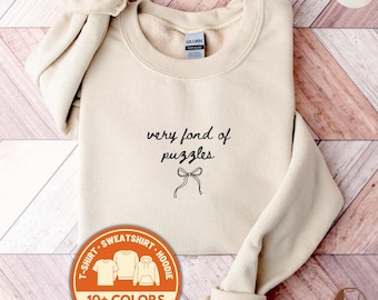 May include: Sand-colored sweatshirt with the text "very fond of puzzles" and a bow graphic. The sweatshirt has a crew neck and long sleeves. A circular graphic shows a t-shirt, sweatshirt, and hoodie, with the text "10+ COLORS" and "FAST SHIPPING".