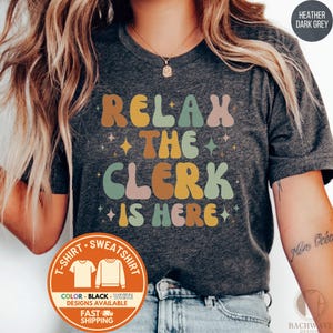 May include: Heather dark grey t-shirt with the text "RELAX THE CLERK IS HERE" in retro-style lettering. The design includes stars and a color palette of brown, pink, yellow, and green. The shirt is available in black and white.