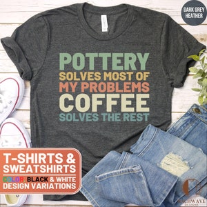 May include: A dark grey heather t-shirt with the text "POTTERY SOLVES MOST OF MY PROBLEMS COFFEE SOLVES THE REST" in a retro font with colorful accents. The text is centered on the shirt. The shirt is displayed on a wooden surface with a pair of jeans and a pair of white sneakers.