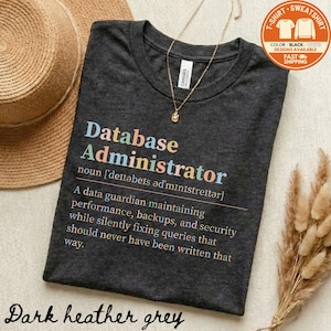 May include: Dark heather grey t-shirt with the text "Database Administrator" in colorful letters. The definition of the term is printed below. A gold necklace and a straw hat are also visible.