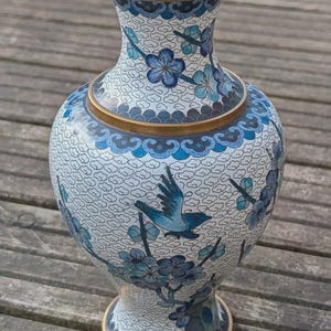 Pode incluir: Um vaso cloisonné com fundo branco e um padrão de nuvens repetitivo, decorado com pássaros azuis e ramos florais azuis. O vaso tem uma borda de metal dourado no topo e uma base de metal dourado a condizer, com bordas recortadas em azul.