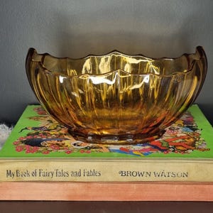 Könnte beinhalten: Eine Vintage-Schale aus bernsteinfarbenem Glas mit gewelltem Rand, die auf einem Stapel Bücher steht. Die Schale hat ein geripptes Design und einen warmen, goldenen Farbton. Die Bücher tragen den Titel "My Book of Fairy Tales and Fables" und "BROWN WATSON".