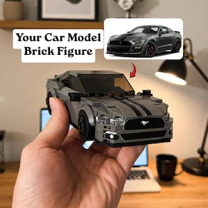 May include: A grey brick car model with black racing stripes. The toy car has black wheels and headlights. The text "Your Car Model Brick Figure" is in a white box in the upper left corner.
