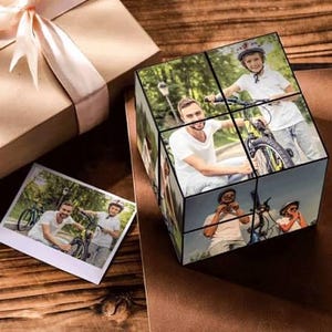 Könnte beinhalten: Ein personalisierter Fotowürfel mit mehreren Bildern eines Mannes und eines Kindes mit Fahrrädern. Ein kleiner Fotoausdruck und eine Geschenkbox mit rosa Schleife sind ebenfalls zu sehen. Der Würfel ist eine einzigartige Geschenkidee.