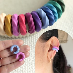 Könnte beinhalten: Eine Sammlung farbenfroher, verdrehter Ohrklemmen aus Polymer Clay in verschiedenen Farbtönen, darunter Blau, Rosa, Lila und Gelb. Das Bild zeigt die Ohrklemmen an einer Hand und an einem Ohr, um das Design und die Tragbarkeit zu veranschaulichen.