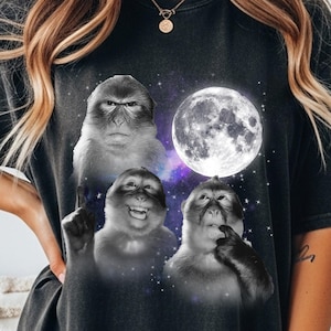 Peut inclure: T-shirt noir avec un motif de trois singes aux expressions variées, sur fond de pleine lune et de ciel étoilé. Les singes sont en niveaux de gris, avec un arrière-plan violet et bleu.