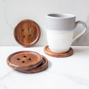 May include: Four wooden coasters with a button design. The coasters are stacked on top of each other. A white mug with a gray band is sitting on top of one of the coasters.
