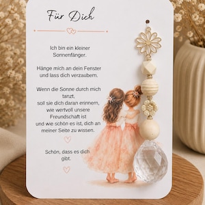 Könnte beinhalten: Eine weiße rechteckige Karte mit einem deutschen Gedicht und einer Illustration von zwei Figuren in pfirsichfarbenen Kleidern. Daran befestigt ist ein Anhänger mit einer goldfarbenen Metallblume, Holzperlen und einem facettierten Kristall.
