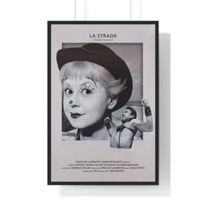 May include: Black and white poster for the film "La Strada" featuring a clown with makeup and a man flexing. The poster includes the film's title, director's name, and cast members. The poster is framed in a black border.