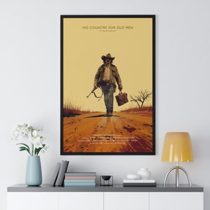 May include: Framed movie poster for "No Country for Old Men" featuring a man in a cowboy hat walking down a dirt road, carrying a rifle and briefcase. The poster has a sepia-toned color palette with text at the top and bottom.