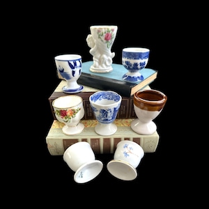 May include: A collection of seven vintage egg cups in various styles and colors, including blue and white floral patterns, brown and white, and a single white egg cup. The egg cups are arranged on a stack of books with the text "The Garden of the Finzi-Continis" visible.