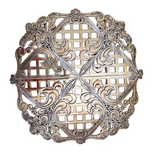 May include: A circular silver-toned metal trivet featuring an intricate openwork design. The pattern consists of four square grid sections separated by ornate, swirling scrollwork motifs that extend toward the scalloped, decorative outer edge.