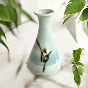 May include: A small, white ceramic vase with a flared opening and a blue and brown painted design. The vase has a delicate, hand-painted floral motif.
