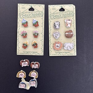 May include: Two Trena's Trinkets cards each holding six buttons with potted plant designs, plus six loose buttons featuring cartoon faces with brown, blonde, and black hair. The cards feature green borders and text reading Trena's Trinkets.