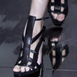 May include: Black leather platform sandals with multiple straps and a high heel. The sandals have an open toe design and a buckle closure. The image shows a close-up of the sandals on a reflective surface. The text in the image says "Runway Gucci S/S 2010 leather".
