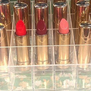Puede incluir: Colección de barras de labios en tubos dorados, expuestas en un organizador transparente y dividido. Se ven tres barras de labios, en tonos rojo, burdeos y melocotón. El organizador está sobre una caja verde con estampado floral.