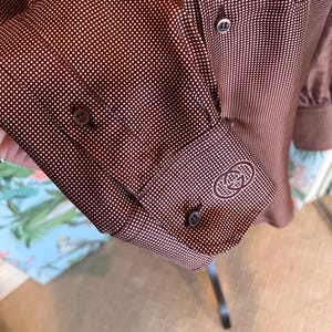 Puede incluir: Una camisa de vestir marrón con estampado de lunares blancos. La camisa tiene cuello abotonado y mangas largas con doble puño. La camisa presenta un pequeño logotipo bordado en el puño izquierdo.