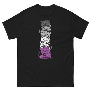 May include: Black t-shirt featuring a stack of cartoon cats in shades of gray, white, and purple. The cats are smiling and stacked on top of each other. The design is centered on the front of the shirt.