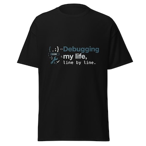 Può includere: Una t-shirt nera a maniche corte con una stampa grafica che riporta il testo "Debugging my life, line by line." Il design include simboli di programmazione come parentesi graffe, un'icona a forma di chiave inglese e una barra di caricamento in blu e bianco.