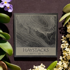 Puede incluir: Un posavasos de pizarra negra con un mapa topográfico de Haystacks, una montaña en los Estados Unidos. El posavasos tiene el texto "Haystacks" y "1.959 FT / 597 M" impreso en él.
