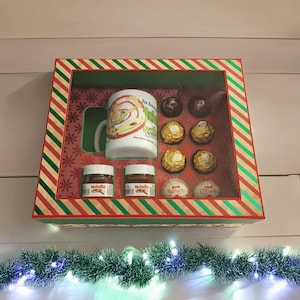 May include: A gift box with a red and green striped border, containing a white mug with a cartoon design, two small jars of Nutella, chocolate candies, and Ferrero Rocher chocolates. The box is decorated with snowflakes.