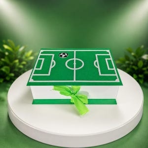 Puede incluir: Una caja de regalo blanca con un diseño de campo de fútbol verde en la tapa. La tapa presenta un gráfico de balón de fútbol. Una cinta verde está atada alrededor de la caja. La caja está sobre una superficie blanca.