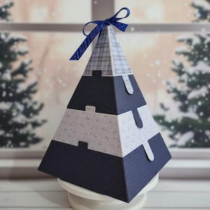 May include: A pyramid-shaped gift box with a navy blue, gray, and white color scheme. The box is made of layered paper with a plaid top and a navy blue ribbon bow. The box is sitting on a white pedestal.