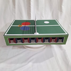 Puede incluir: Una caja con temática de tenis de mesa verde y blanca con una red, paletas y una pelota. La caja tiene ocho compartimentos numerados. Las paletas son rojas y azules. Los números son en fuente plateada.