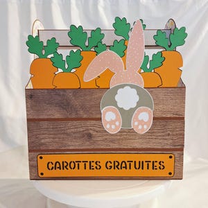 Peut inclure: Une boîte en bois avec un design de lapin et de carottes. La boîte présente des carottes oranges avec des fanes vertes et un lapin rose et blanc à pois. Le texte "CARROTTES GRATUITES" est sur un panneau orange à l'avant de la boîte.